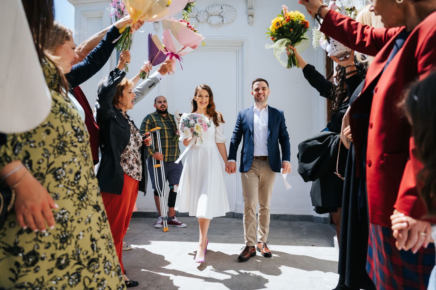 Newlywed couple walks through floral arch as guests celebrate outside a white chapel on a sunny wedding day.