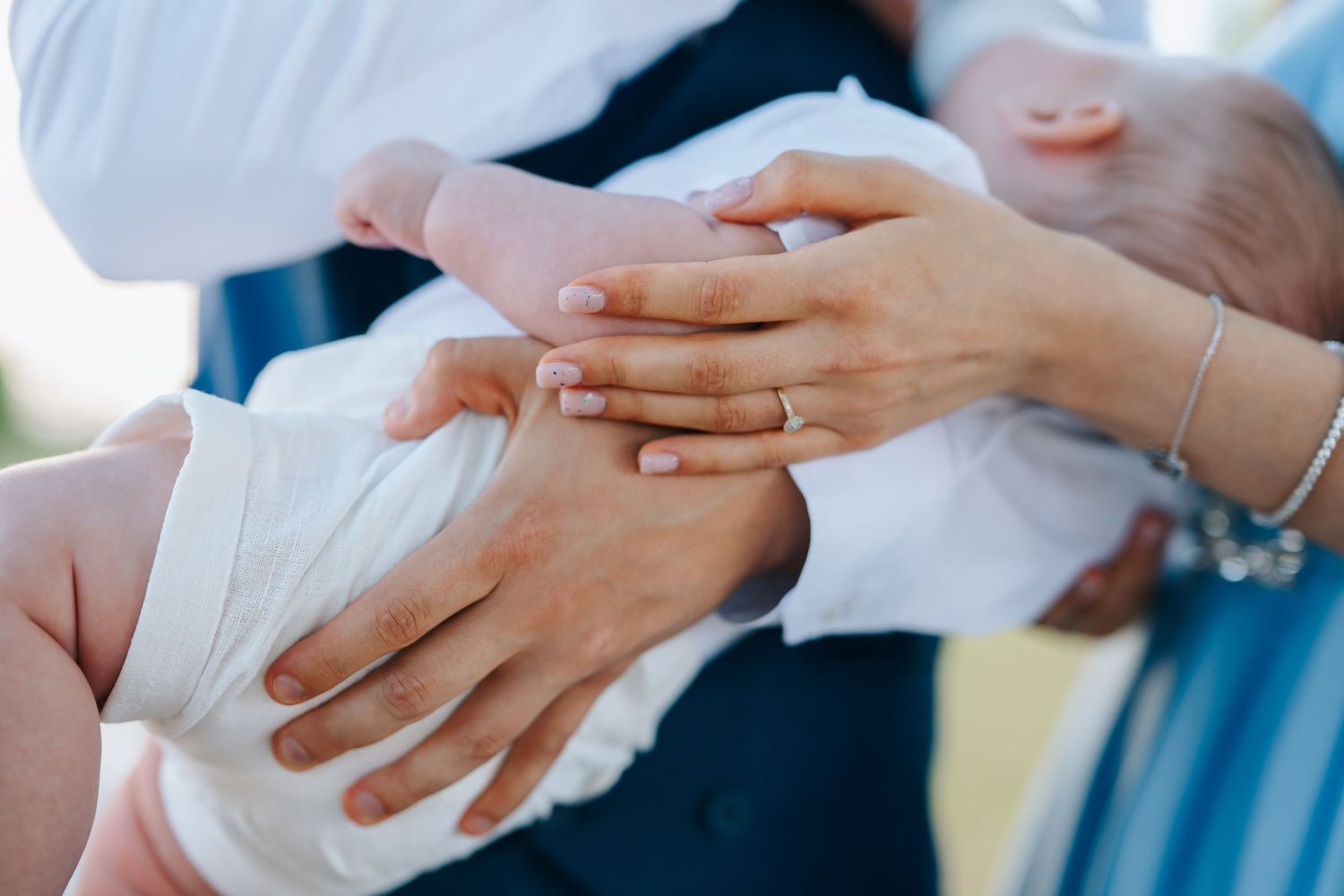 Married couple holding newborn baby, showing wedding rings and a delicate bracelet at an outdoor ceremony.