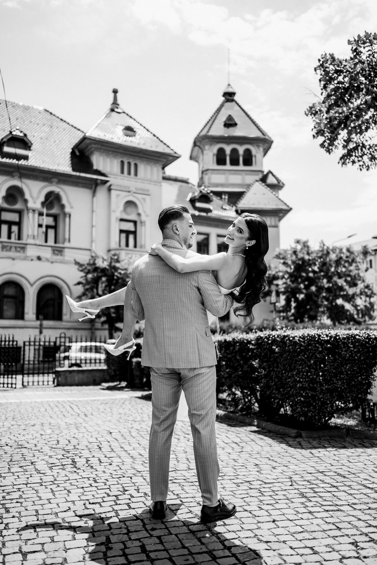 Groom lifts smiling bride outdoors in black and white photo with historic European building in background.