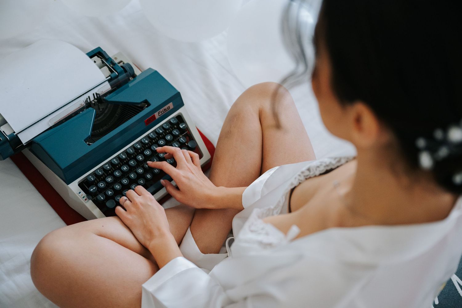 Woman in white robe typing on a vintage teal typewriter while sitting cross-legged on a bed.