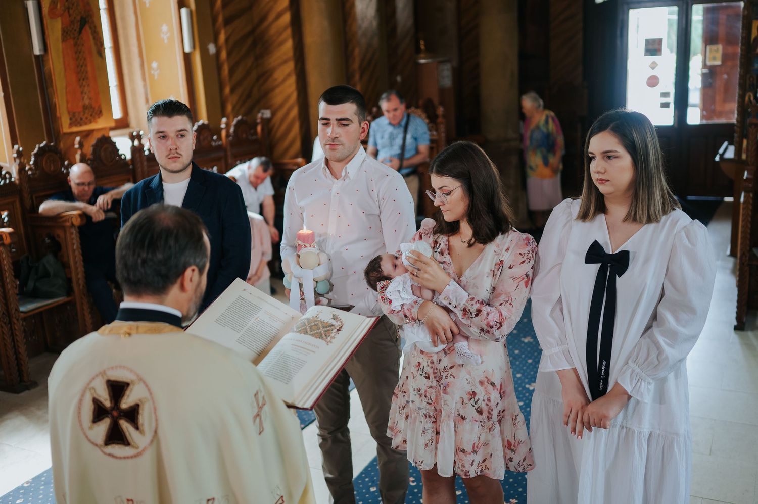 A priest performs a baptism ceremony for a baby held by a woman in a floral dress inside a church.