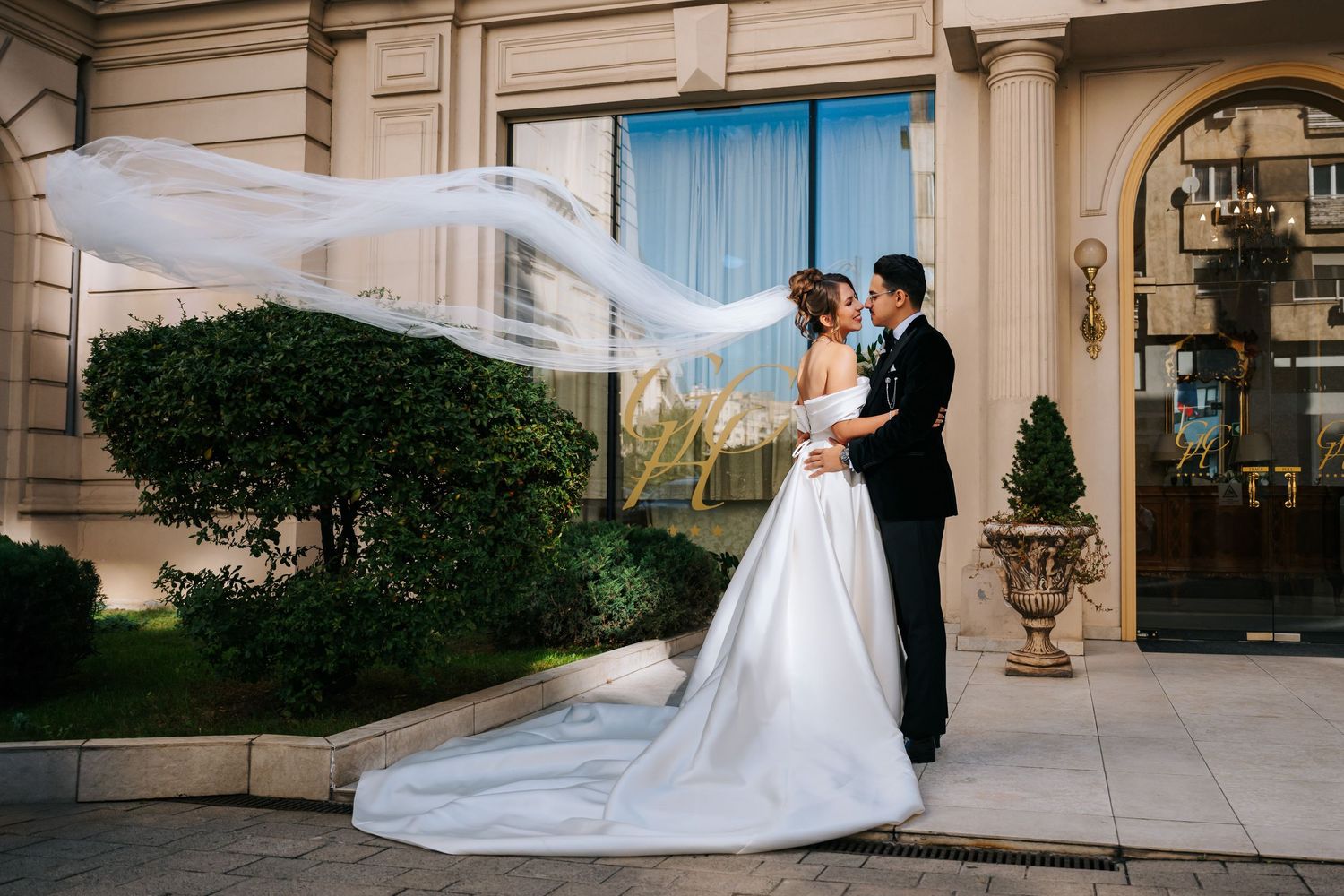Bride and groom embrace outside elegant venue as wedding veil flows dramatically in the wind.