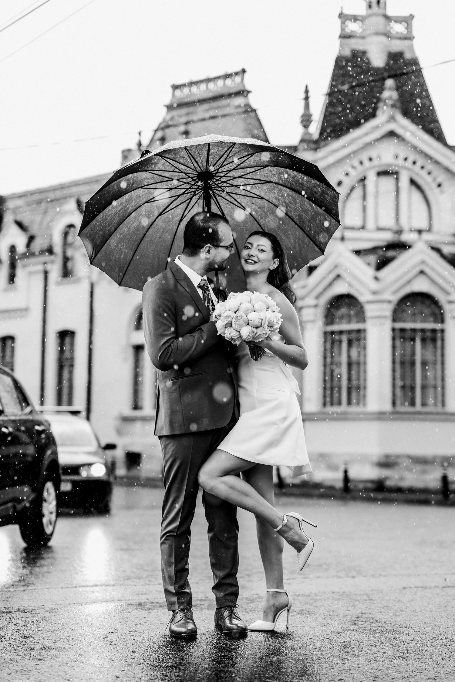 Black and white photo of newlyweds sharing romantic moment under umbrella in rainy street.