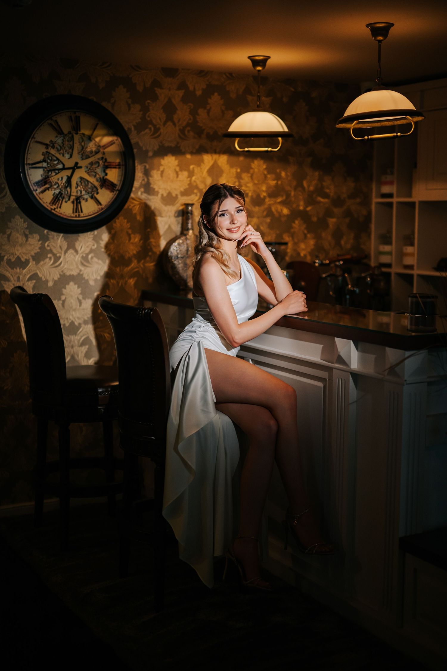 Elegant woman in white dress sitting at a bar counter in a dimly lit room with vintage decor and pendant lights.
