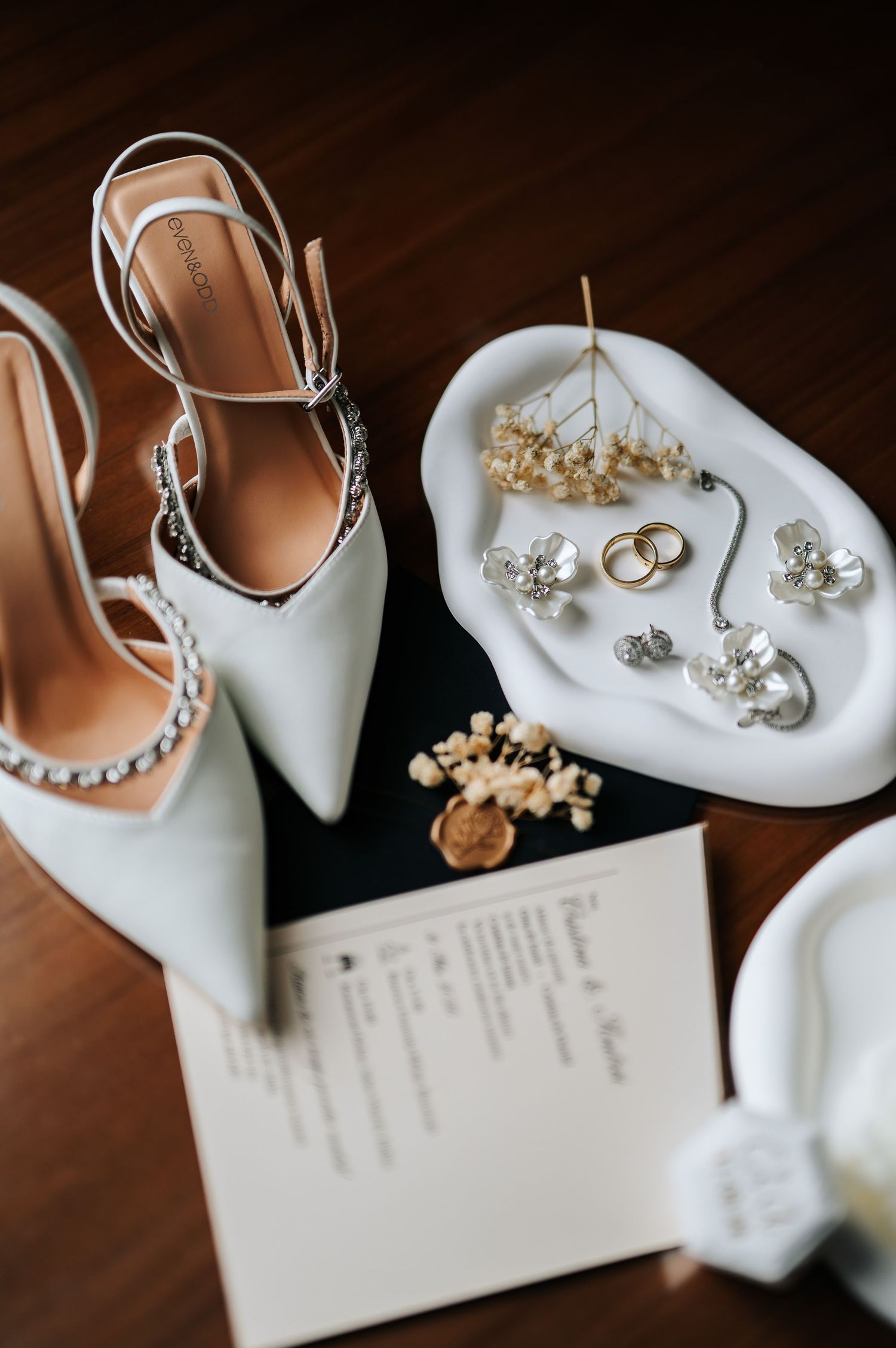 Elegant wedding flat lay featuring white heels, gold rings, pearl jewelry, and invitation on dark wood surface.