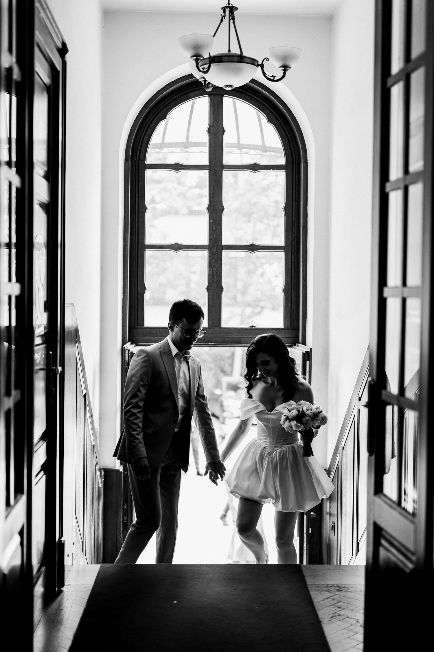 Black and white silhouette of couple holding hands before arched window on staircase.