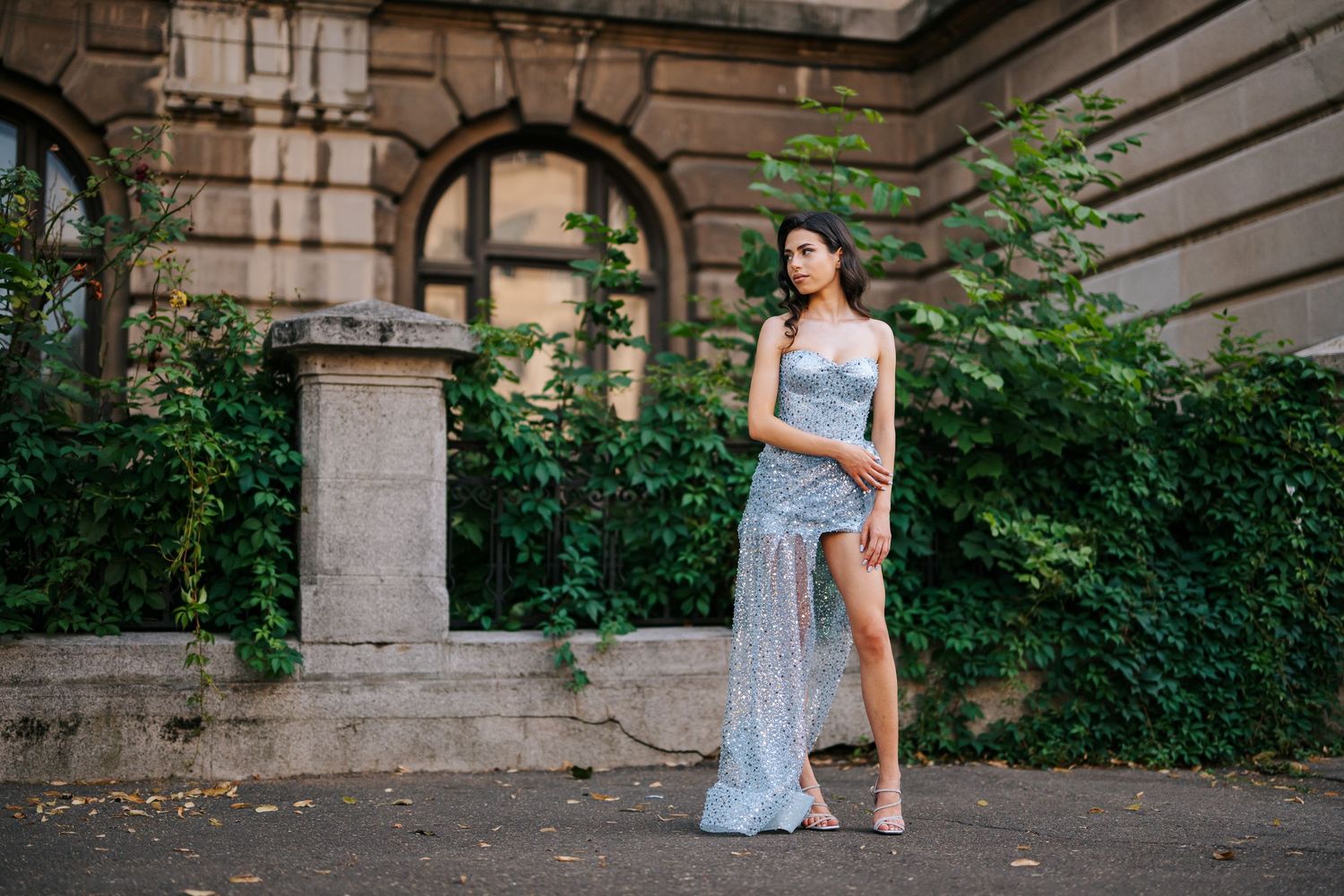 Woman in elegant silver sequin gown poses outdoors near stone building with lush green ivy backdrop.