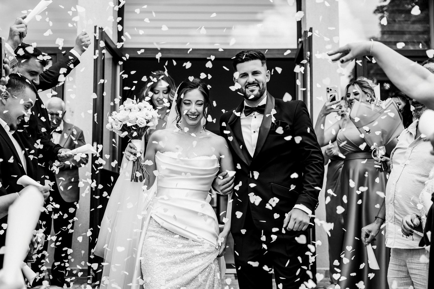 Black and white photo of a joyful bride and groom walking through confetti shower surrounded by cheering wedding guests.