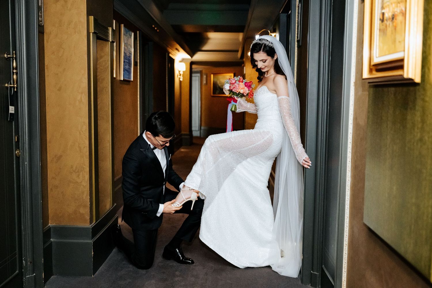 Groom kneels to adjust bride's shoe in elegant hotel corridor on wedding day.