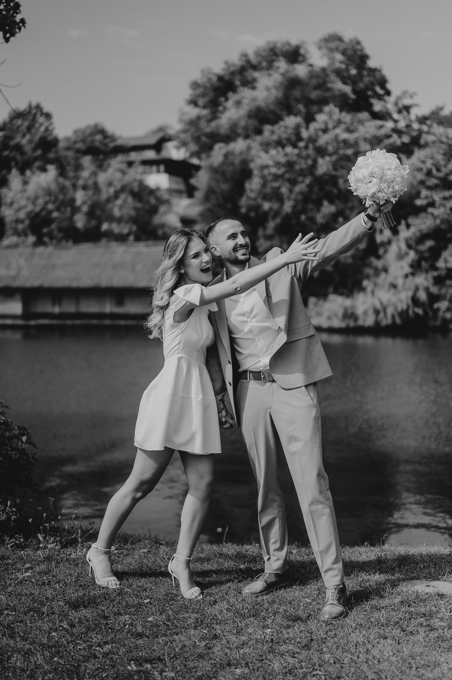 Happy couple celebrating outdoors near lake in black and white photo holding white bouquet.