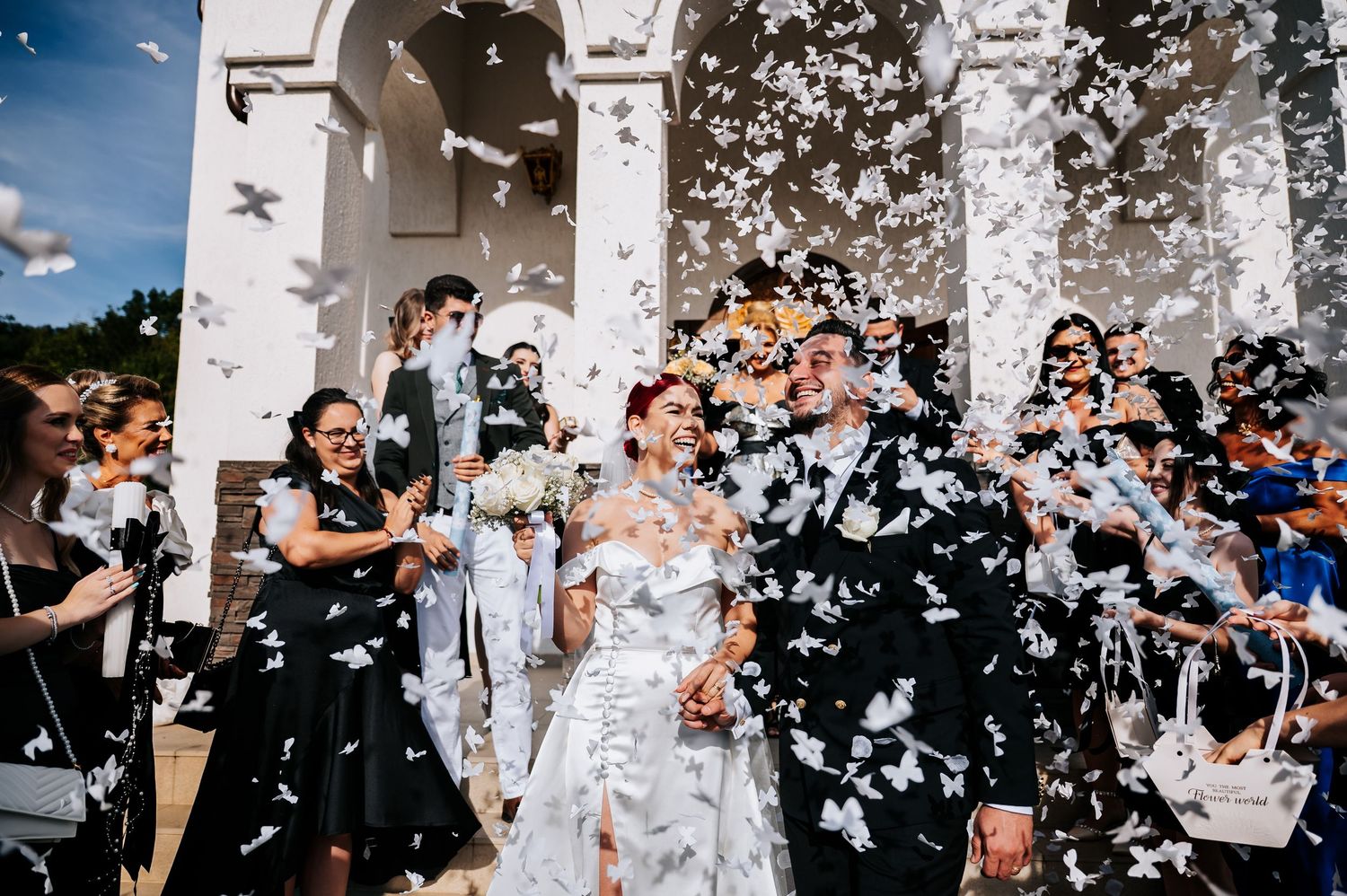 Bride and groom exit church ceremony surrounded by guests throwing white butterfly confetti outdoors.