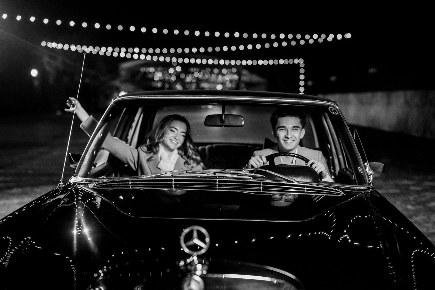 Couple smiling in a vintage Mercedes-Benz car at night with string lights in black and white photo.