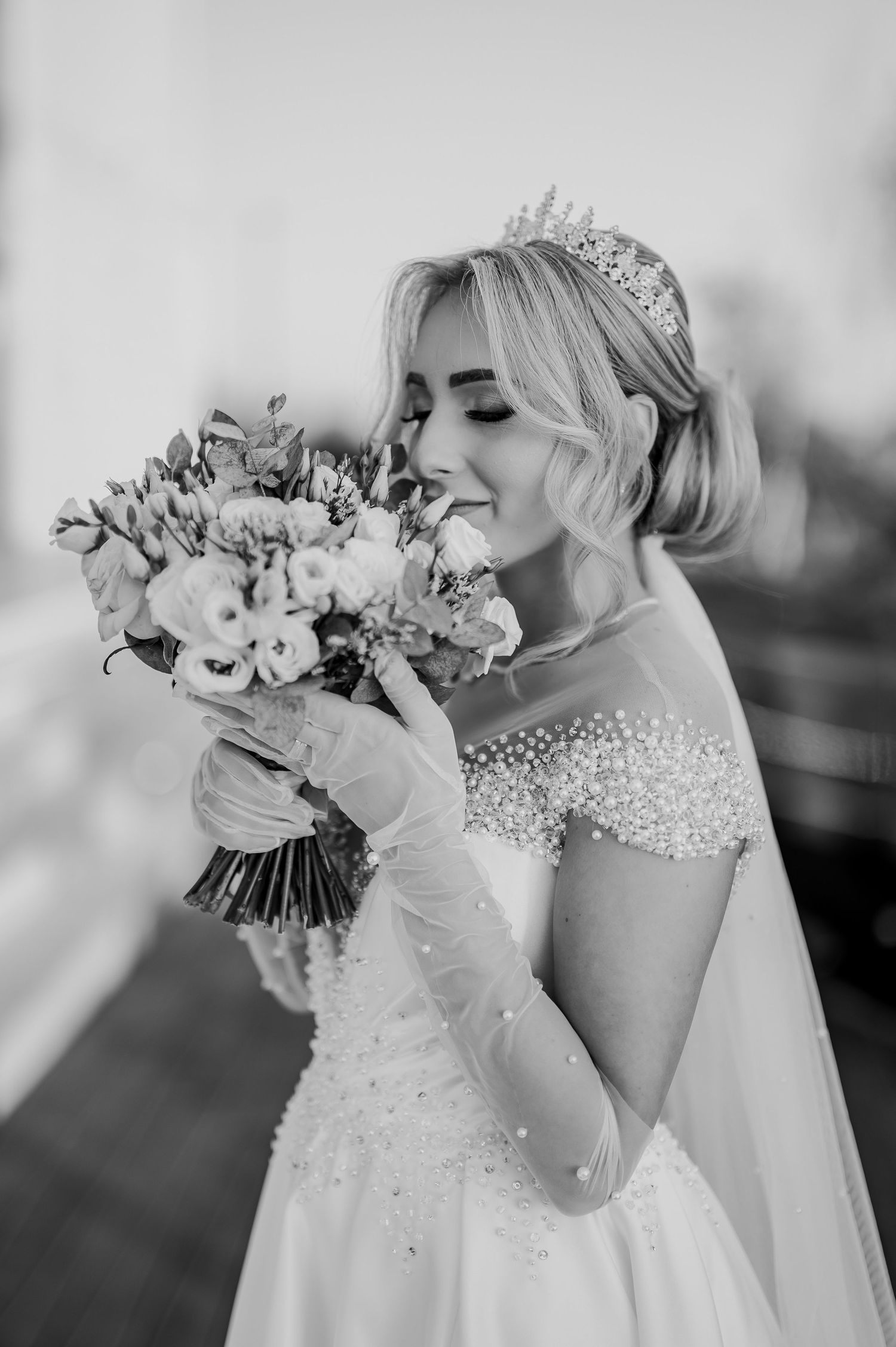Black and white photo of a smiling bride in a pearl gown and tiara, holding a bouquet of flowers.