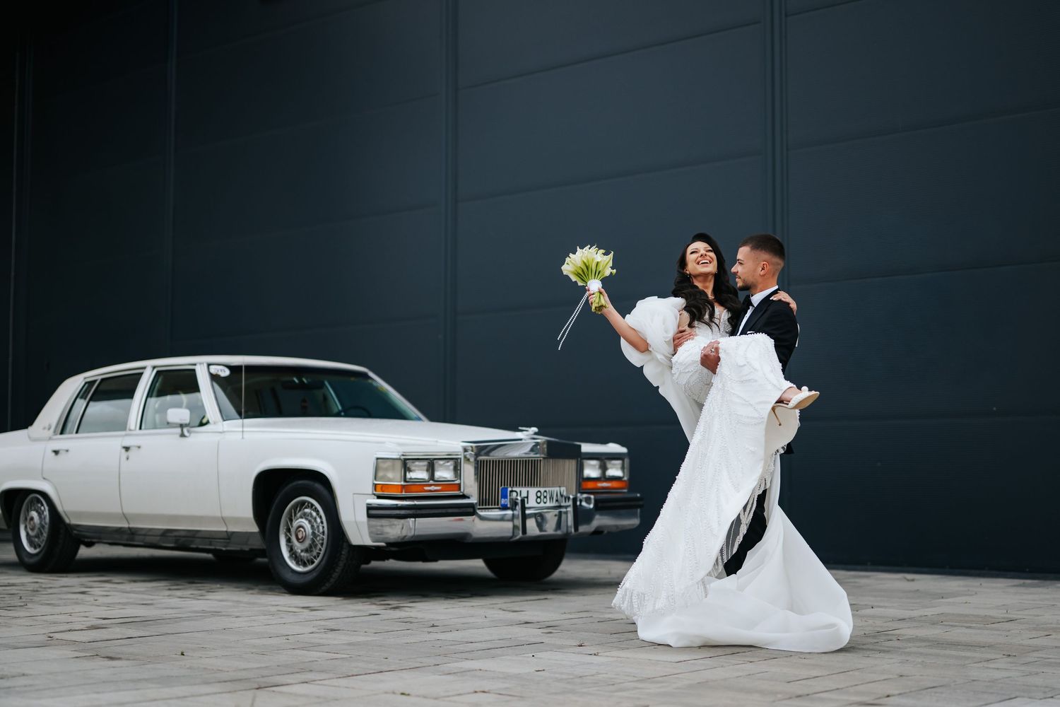 Groom dips bride in white gown holding yellow bouquet beside vintage white Cadillac against dark wall.