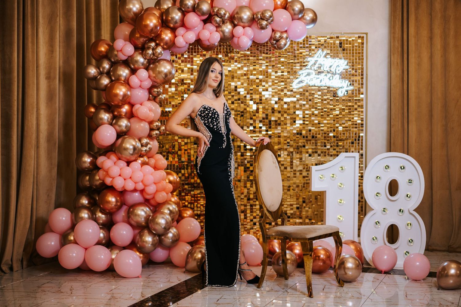 Young woman in elegant black gown poses at an 18th birthday party with pink and gold balloon decorations.