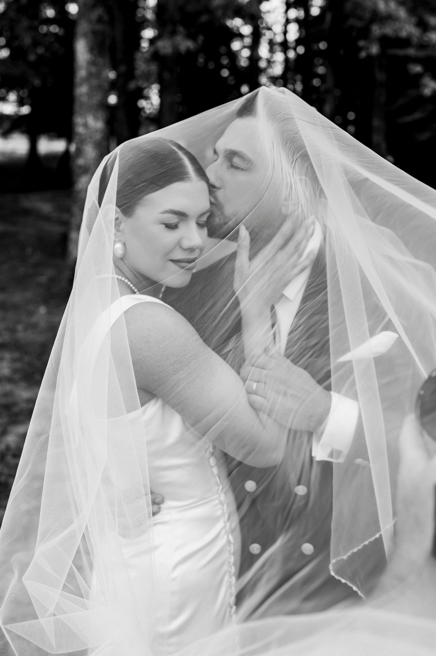 Black and white photo of bride and groom sharing a romantic kiss under a flowing wedding veil outdoors.