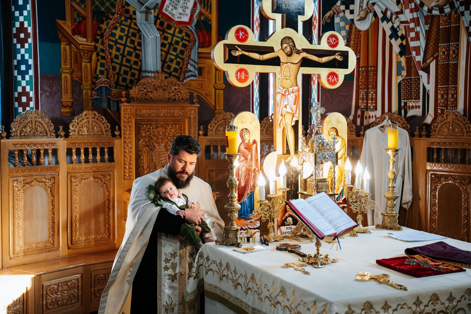 Orthodox priest holding infant near candlelit altar with ornate iconostasis during baptism ceremony.