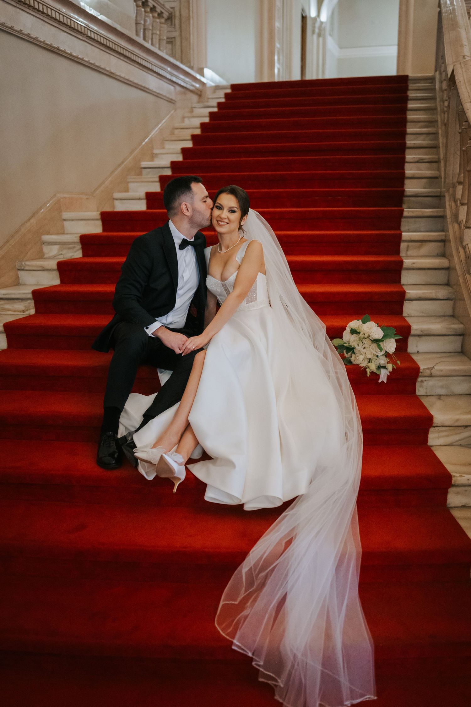 Bride and groom sitting on grand staircase with red carpet, sharing a kiss in elegant wedding attire.
