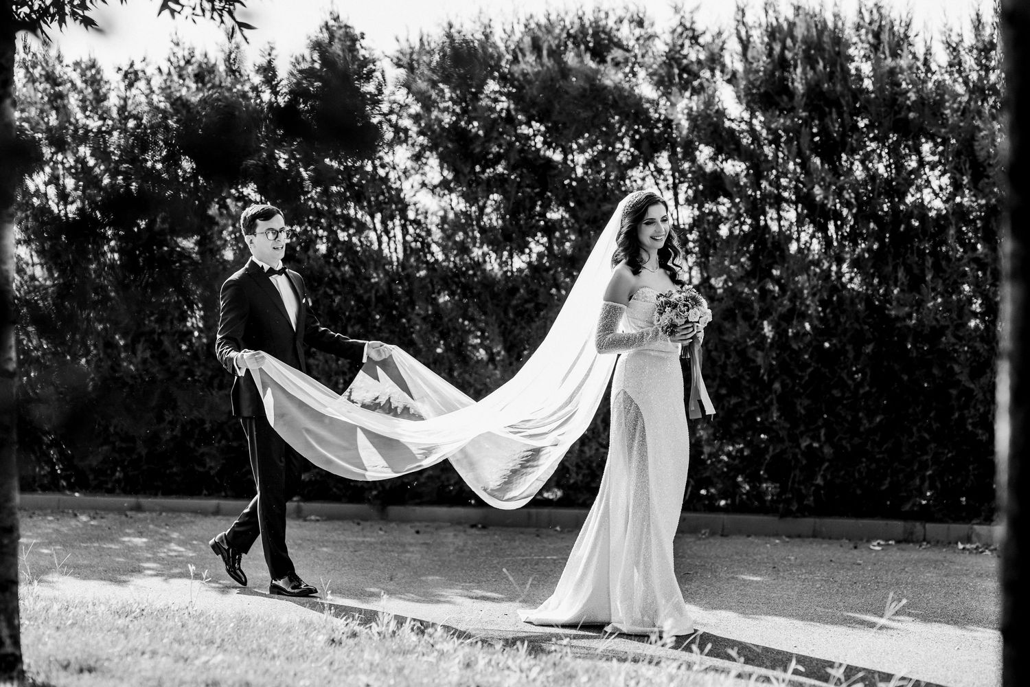 Black and white wedding photo of groom holding bride's flowing veil outdoors with lush green trees in background.
