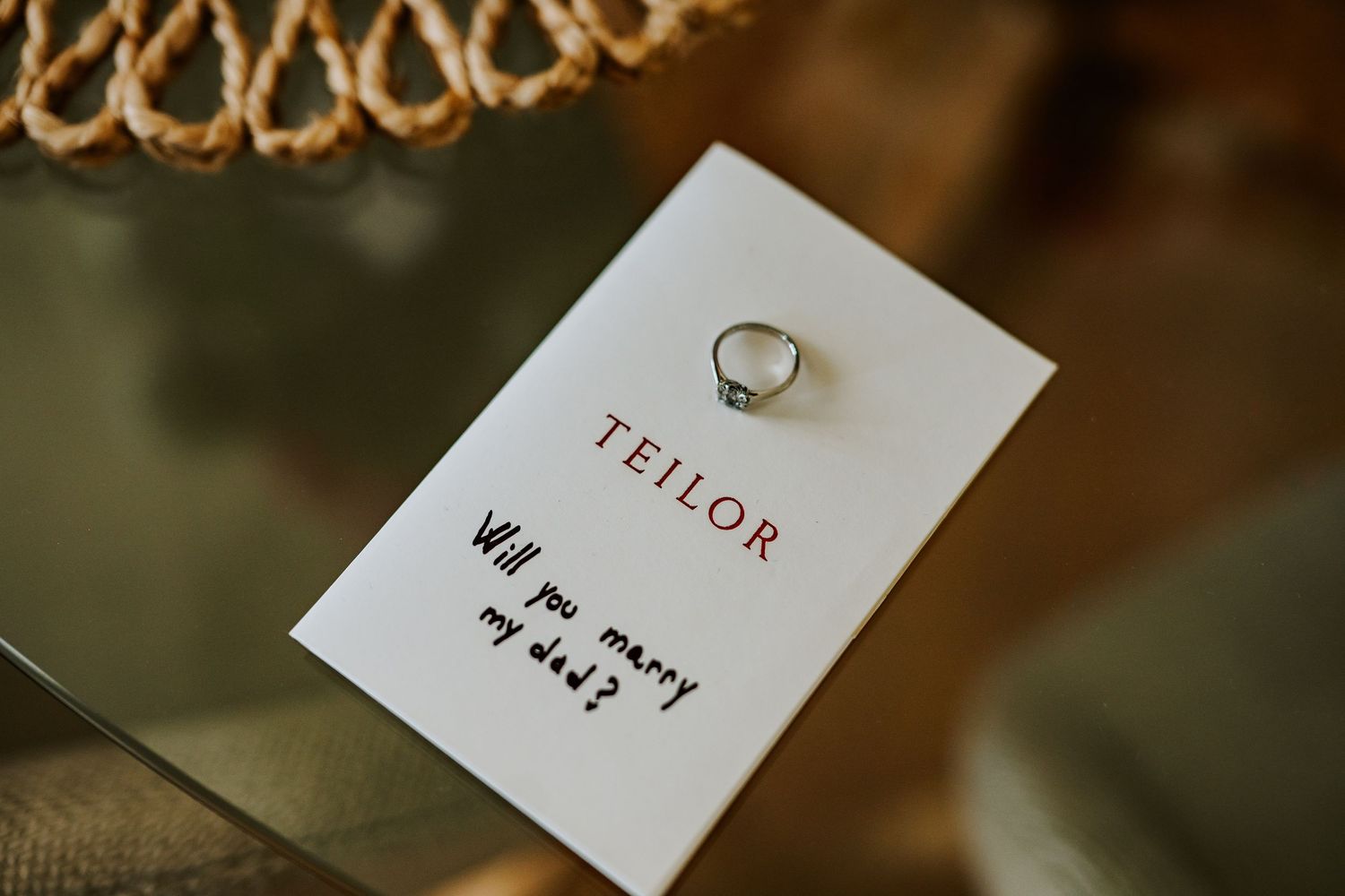 A Teilor engagement ring on a card reading Will you marry me? displayed on a glass surface.