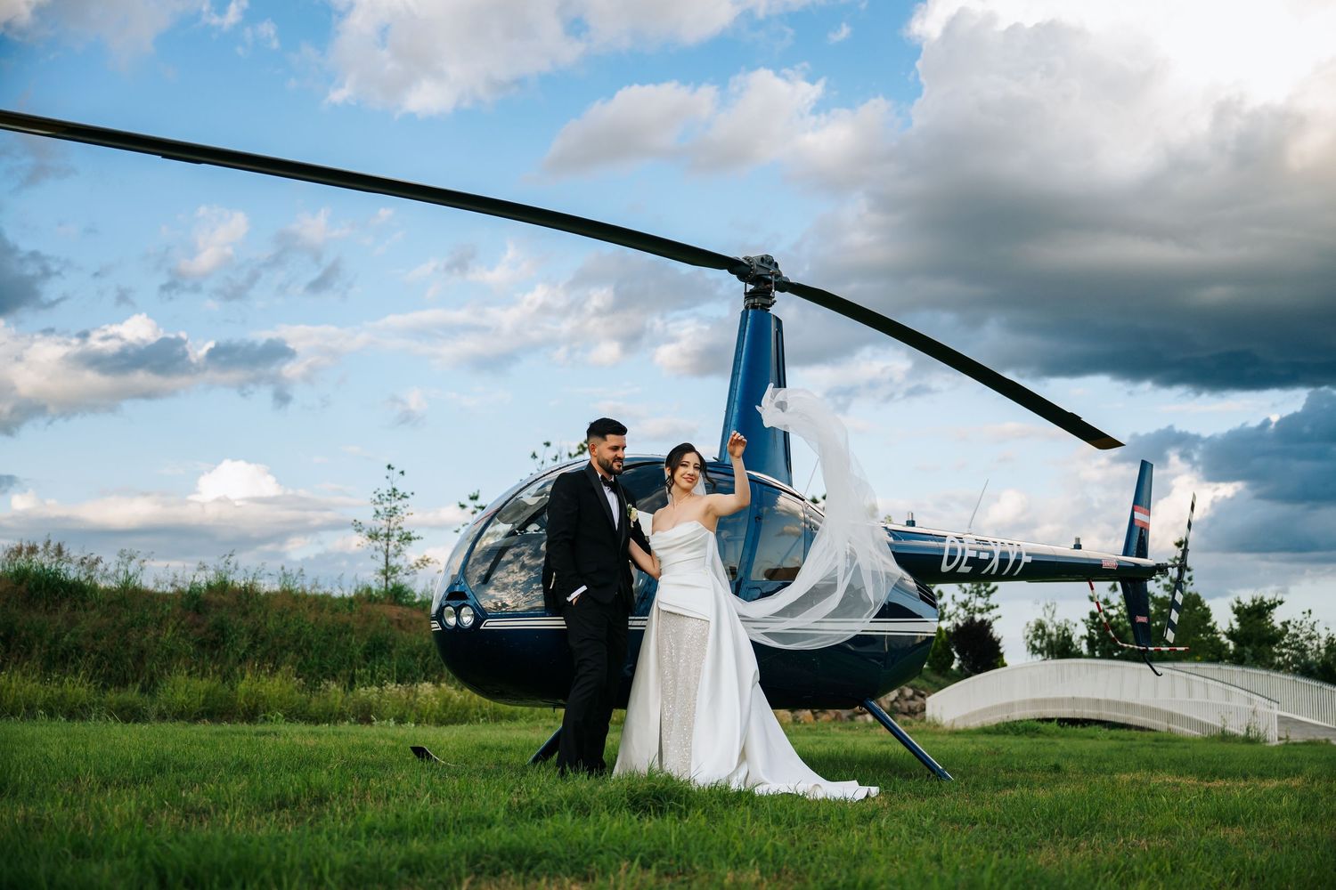 Bride and groom posing in front of a blue helicopter on a green field with dramatic cloudy sky.