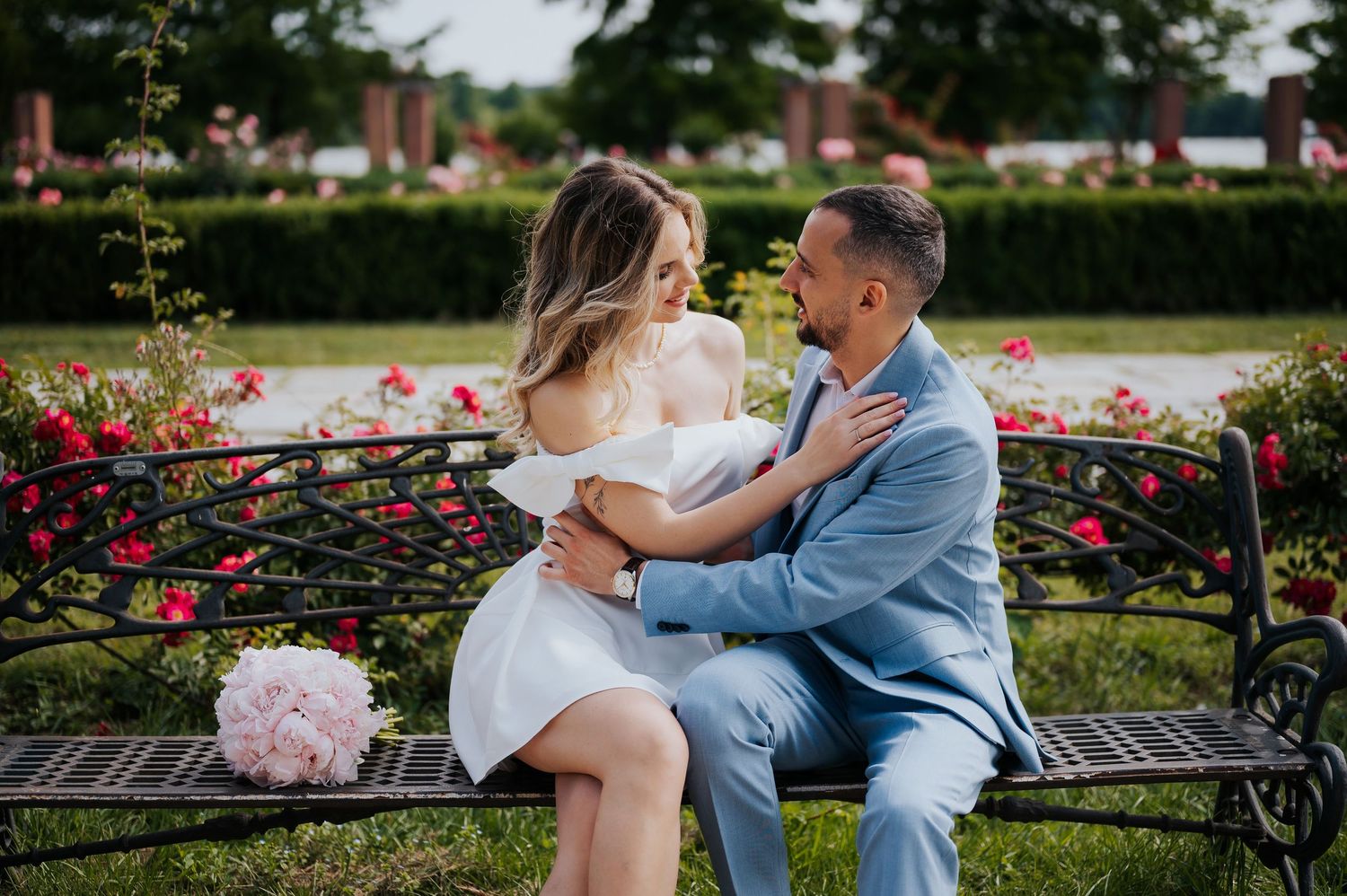 Couple in wedding attire sitting on a bench surrounded by roses, sharing a romantic moment outdoors.