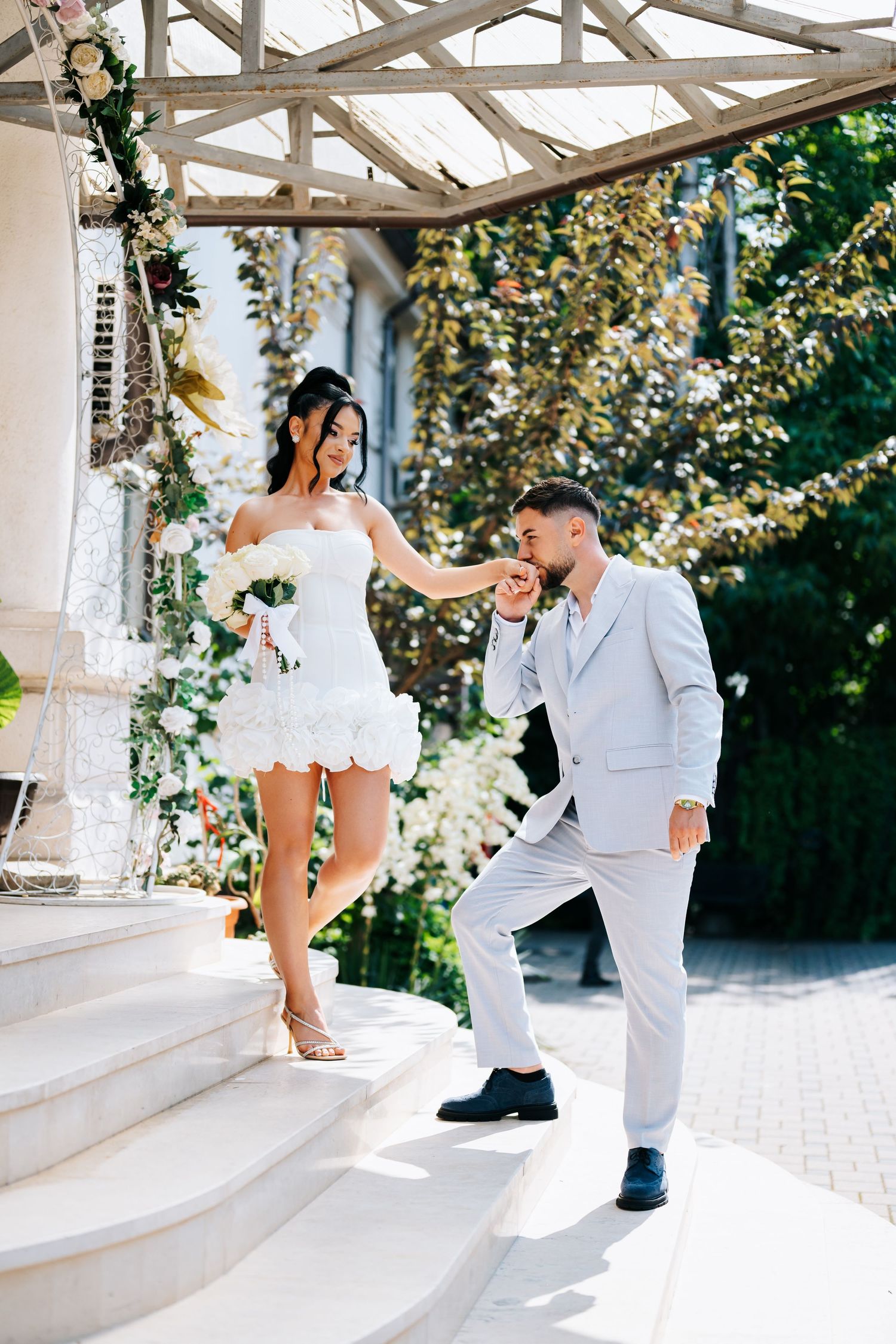 Groom gallantly helps bride in short white feathered dress descend outdoor steps at sunny wedding venue.