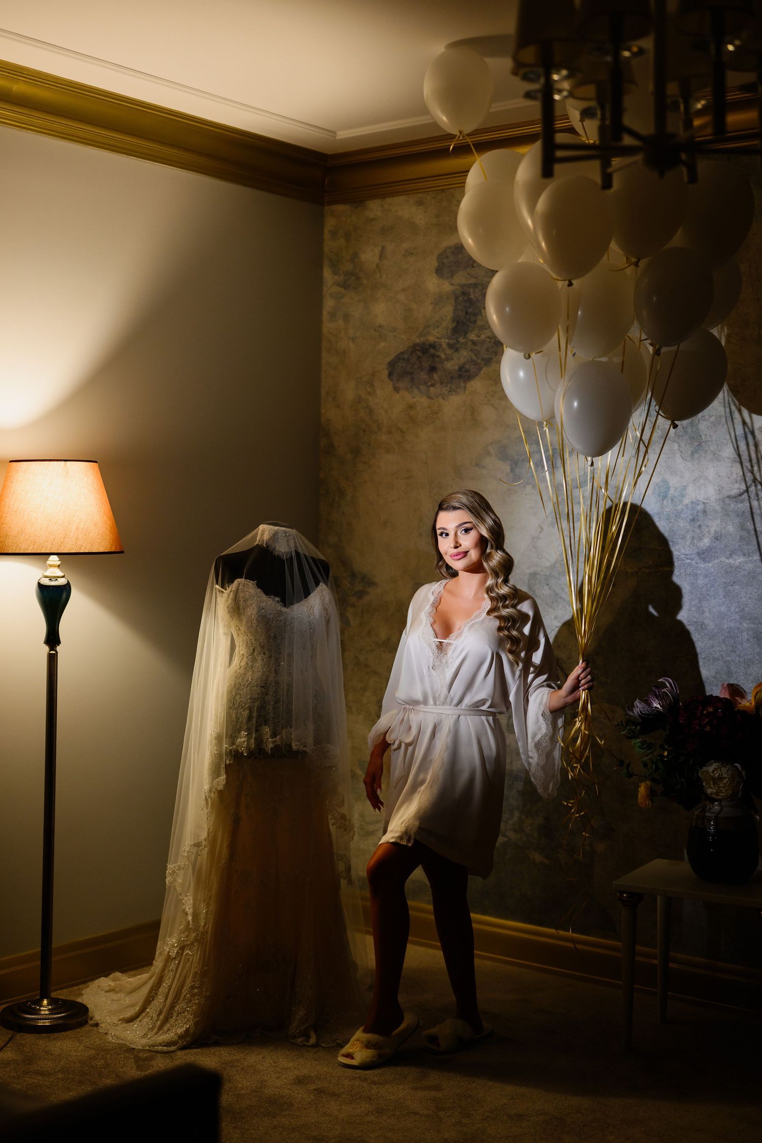 Bride in white robe holding balloons beside wedding dress on mannequin in elegant hotel room.