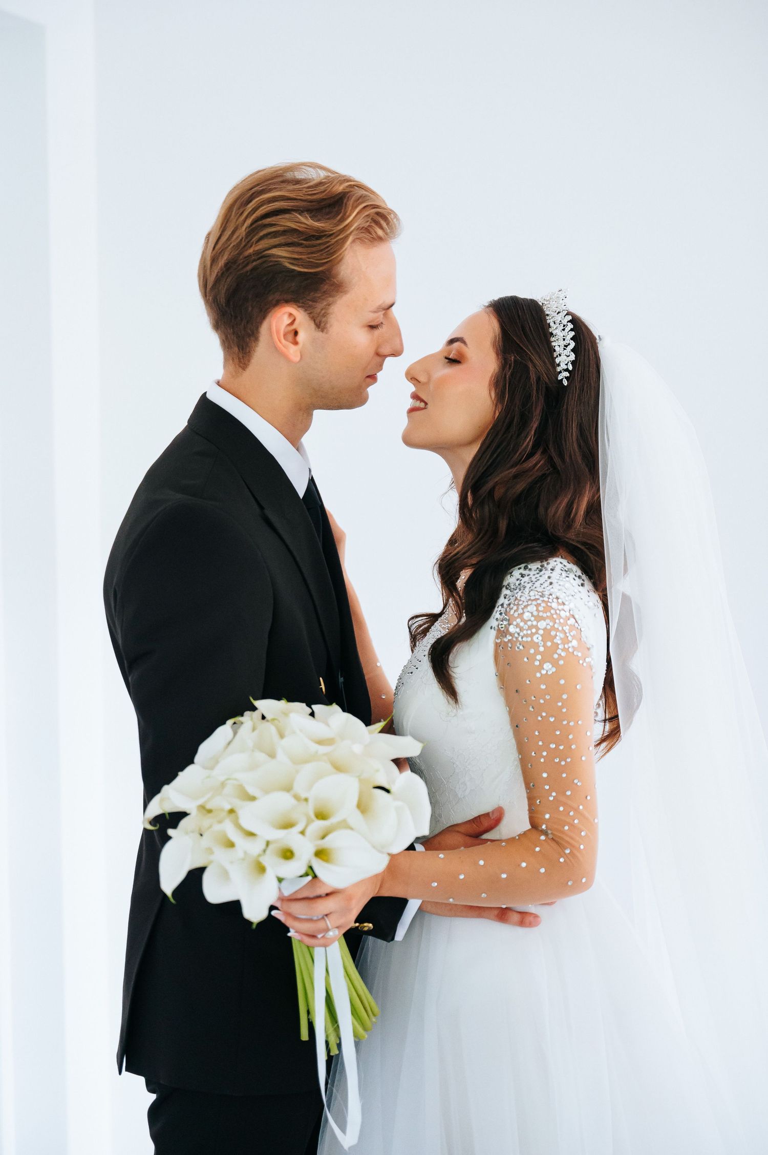 Bride and groom share romantic almost-kiss holding white calla lily bouquet in bright studio.