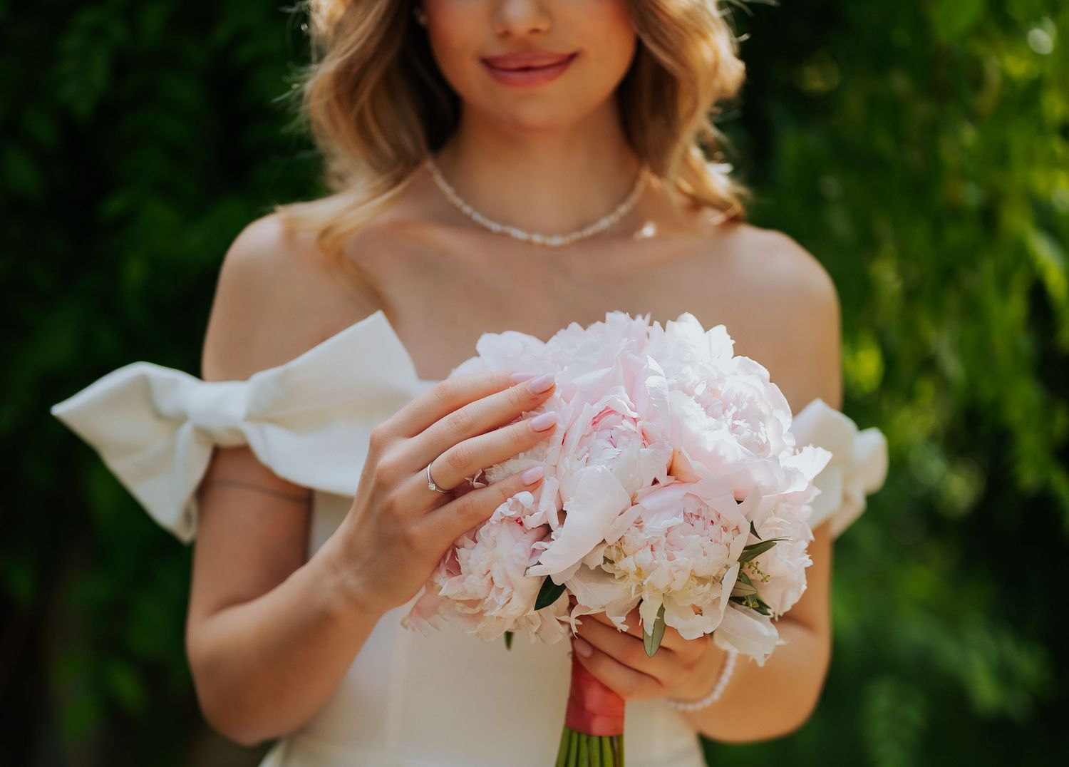 Bride in off-shoulder white dress holding soft pink peony and hydrangea bouquet with diamond ring.