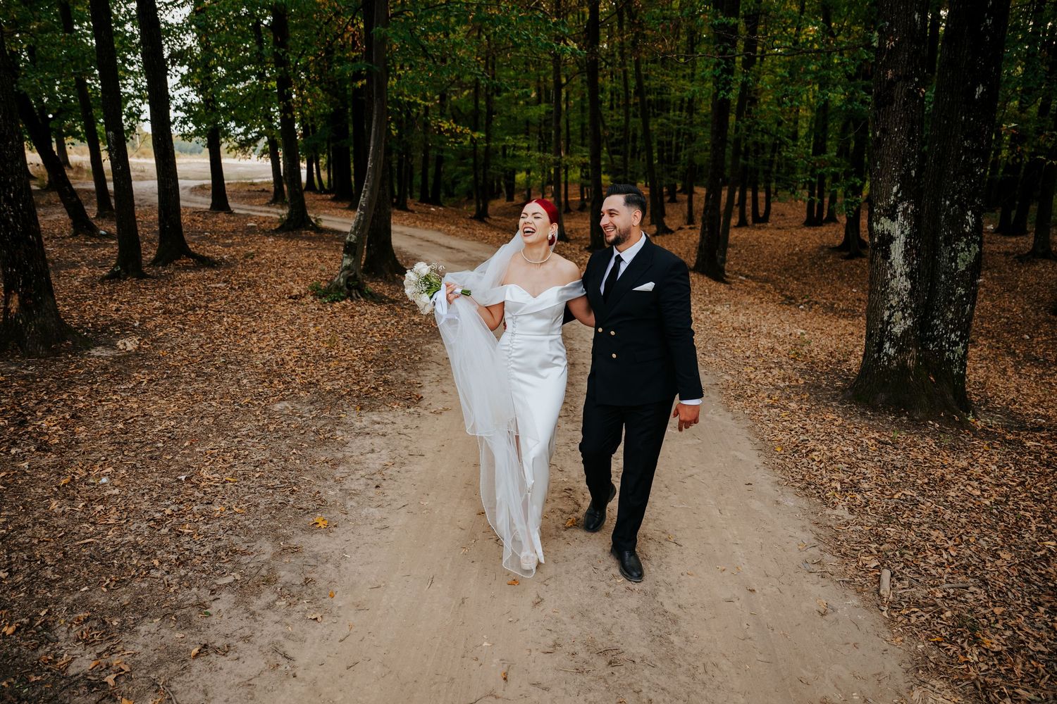 A bride and groom walk hand-in-hand along a sandy forest path surrounded by tall trees and autumn leaves.