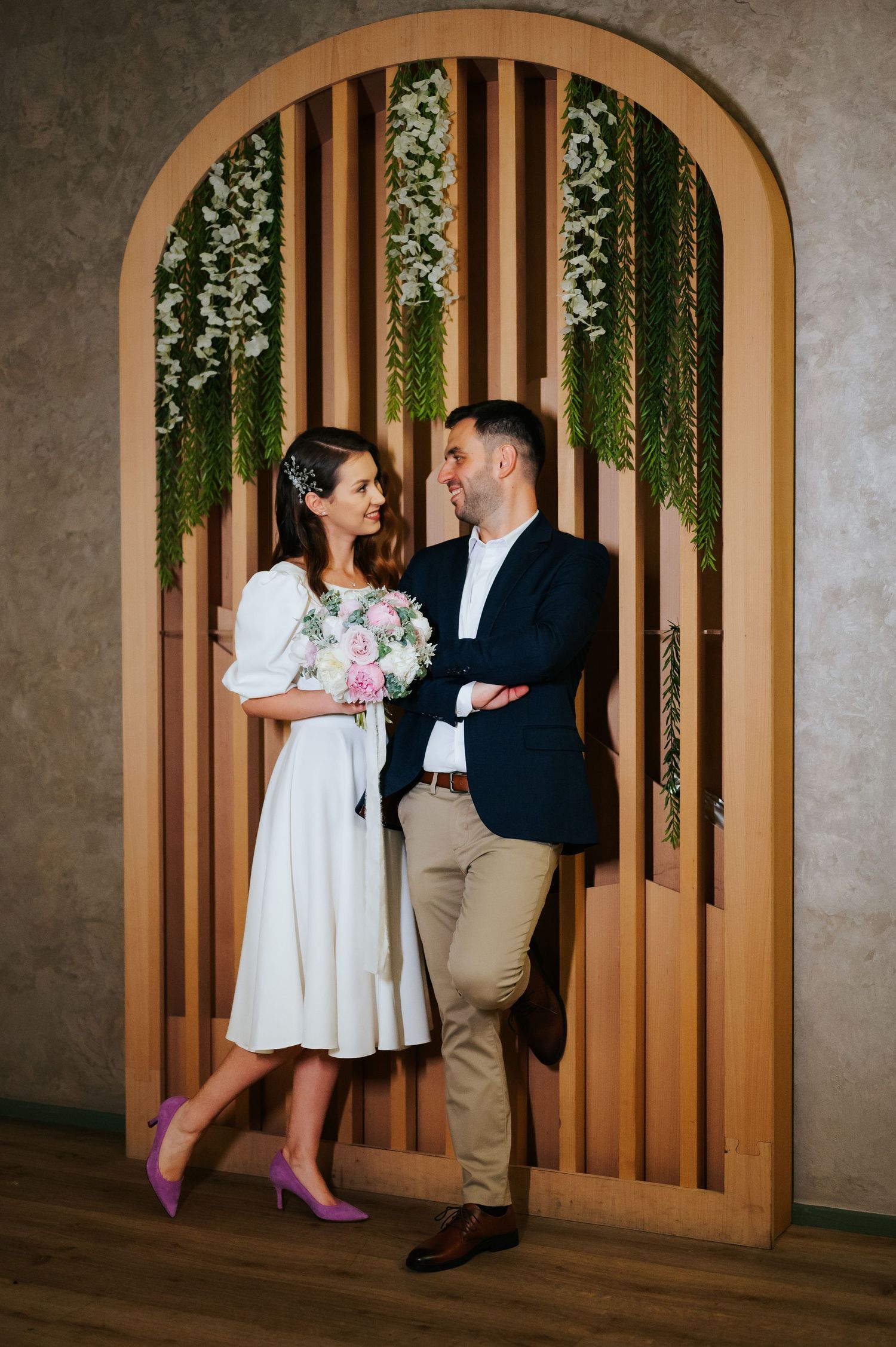 Couple in wedding attire standing before a wooden arch with greenery, bride holding pink bouquet, groom in navy blazer.