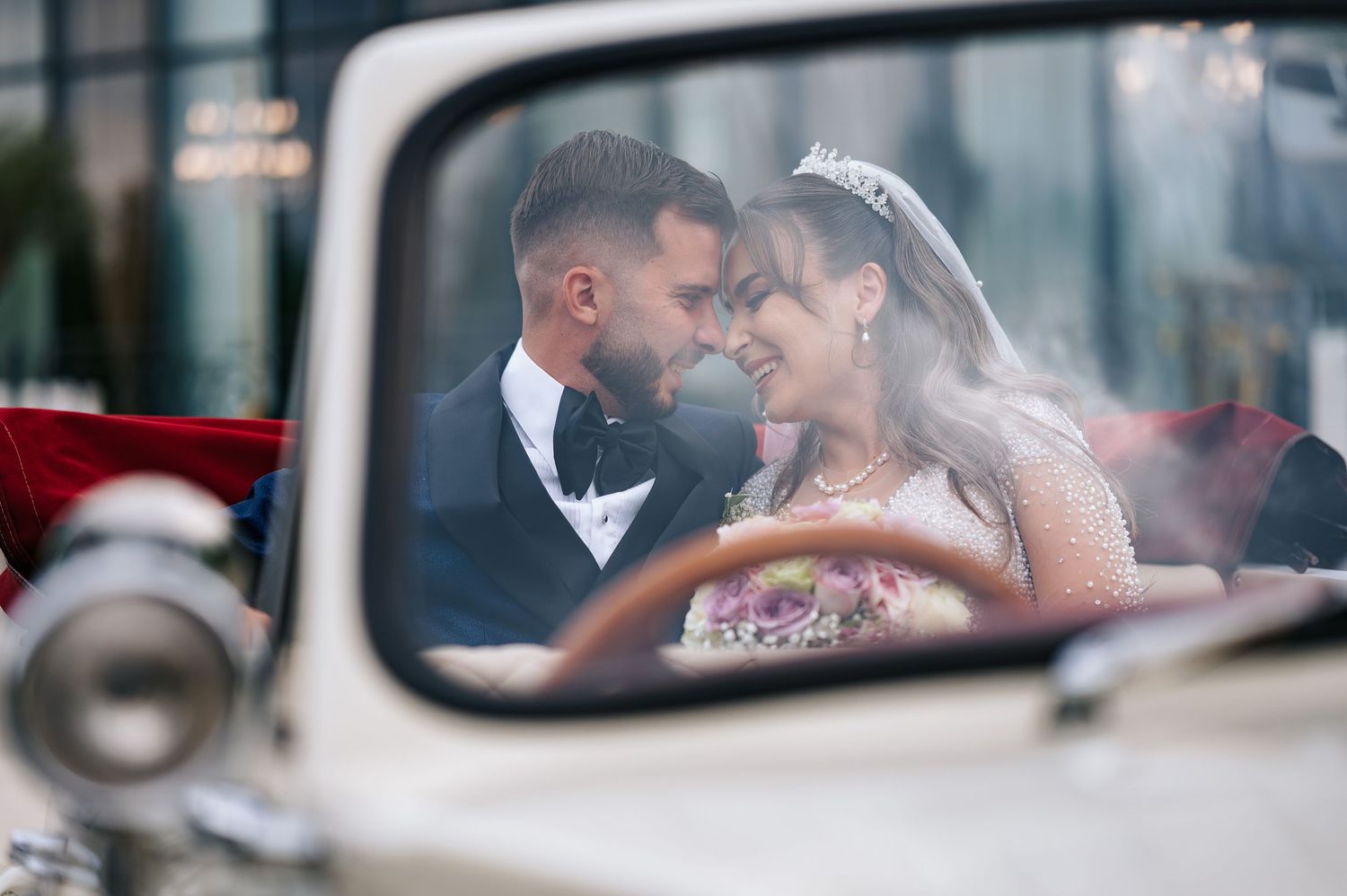 Bride and groom share an intimate moment inside a classic vintage car, seen through the windshield.