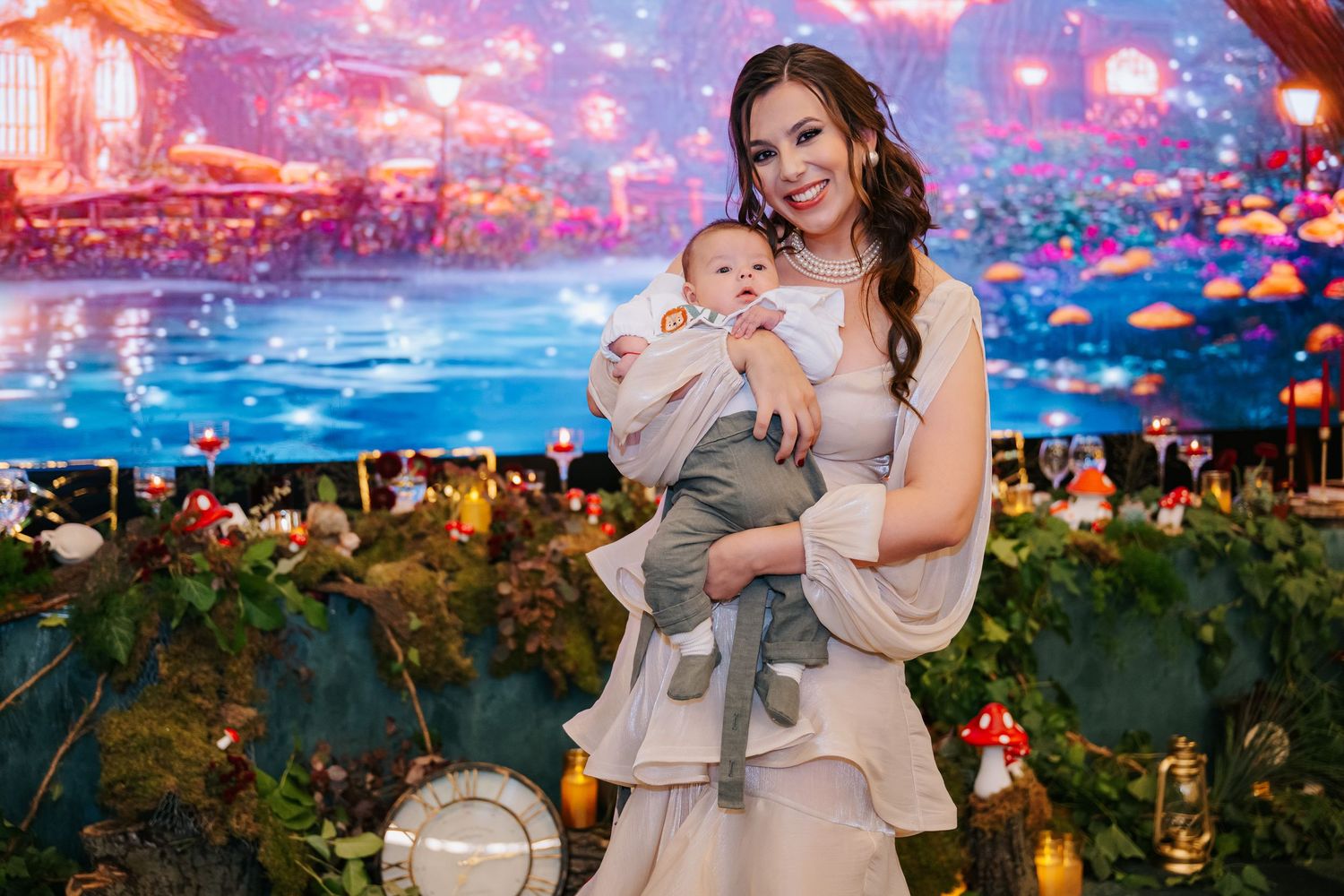 Smiling mother in white dress holding baby boy at magical fantasy-themed birthday party backdrop.