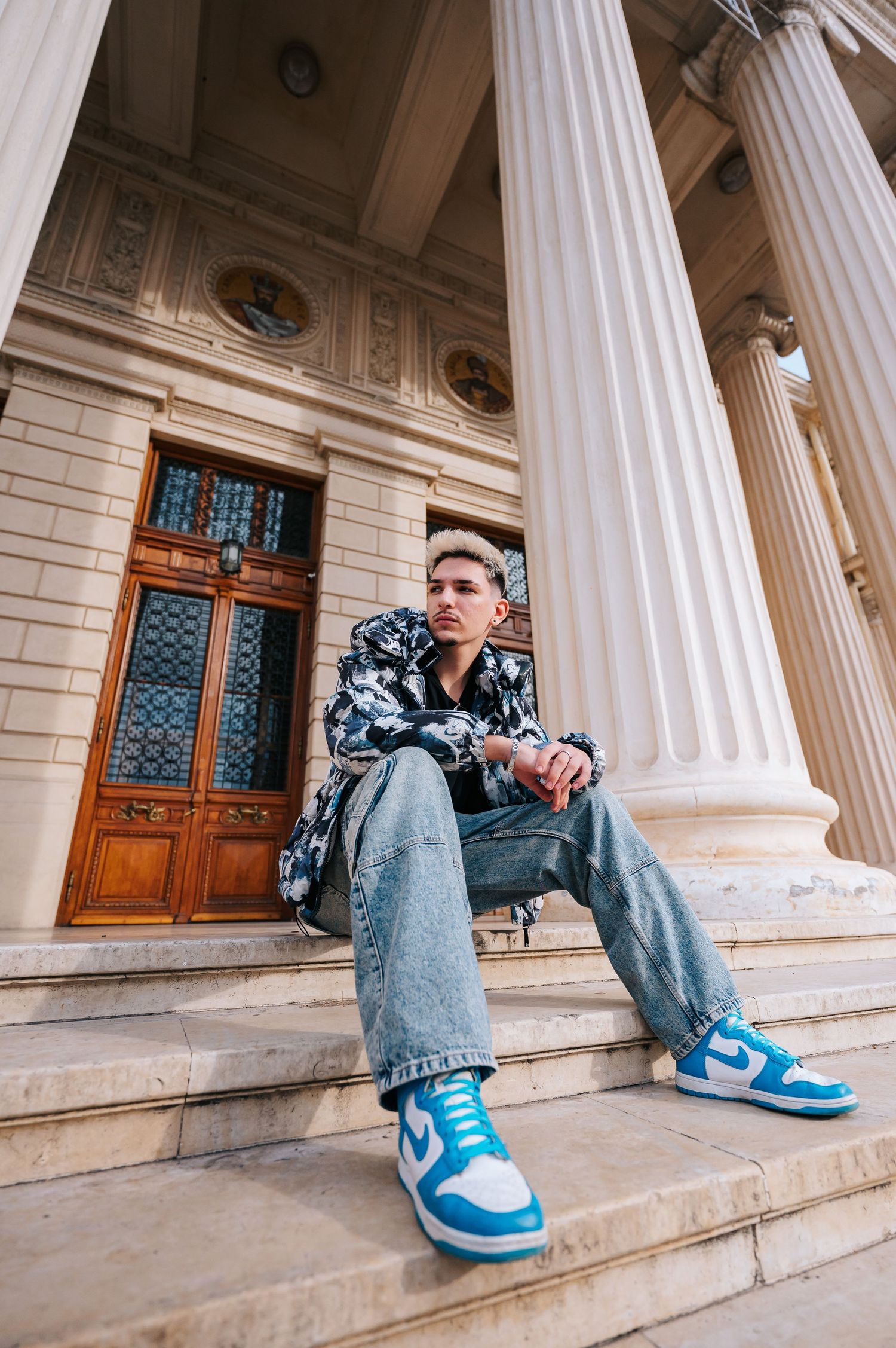 Young man in blue Nike sneakers and jeans sitting on steps of a neoclassical building with white columns.