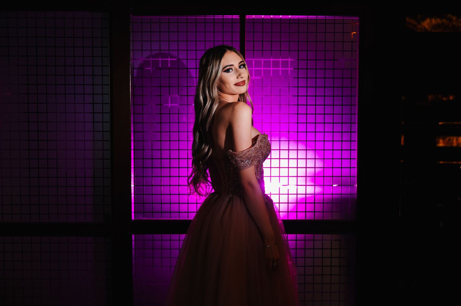 Woman in elegant off-shoulder gown posing against glowing pink grid backdrop in dramatic portrait photography.