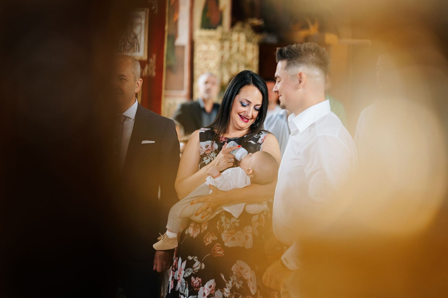 A woman in a floral dress holds a baby during a christening ceremony, with a man beside her in a church.