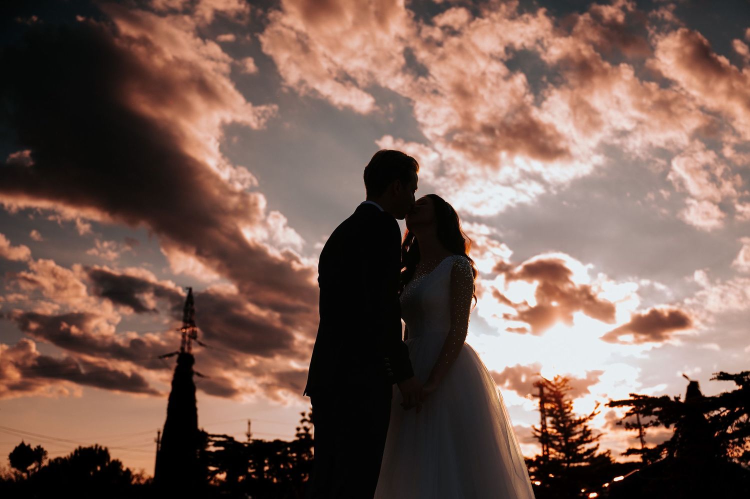 Silhouette of a bride and groom kissing against a dramatic sunset sky with glowing clouds at golden hour.