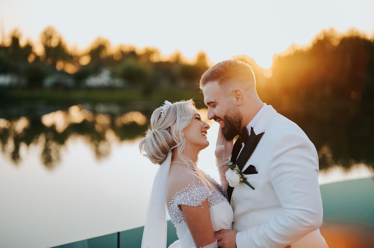 Bride and groom share a romantic moment at golden hour by a lake, sun glowing warmly behind them.