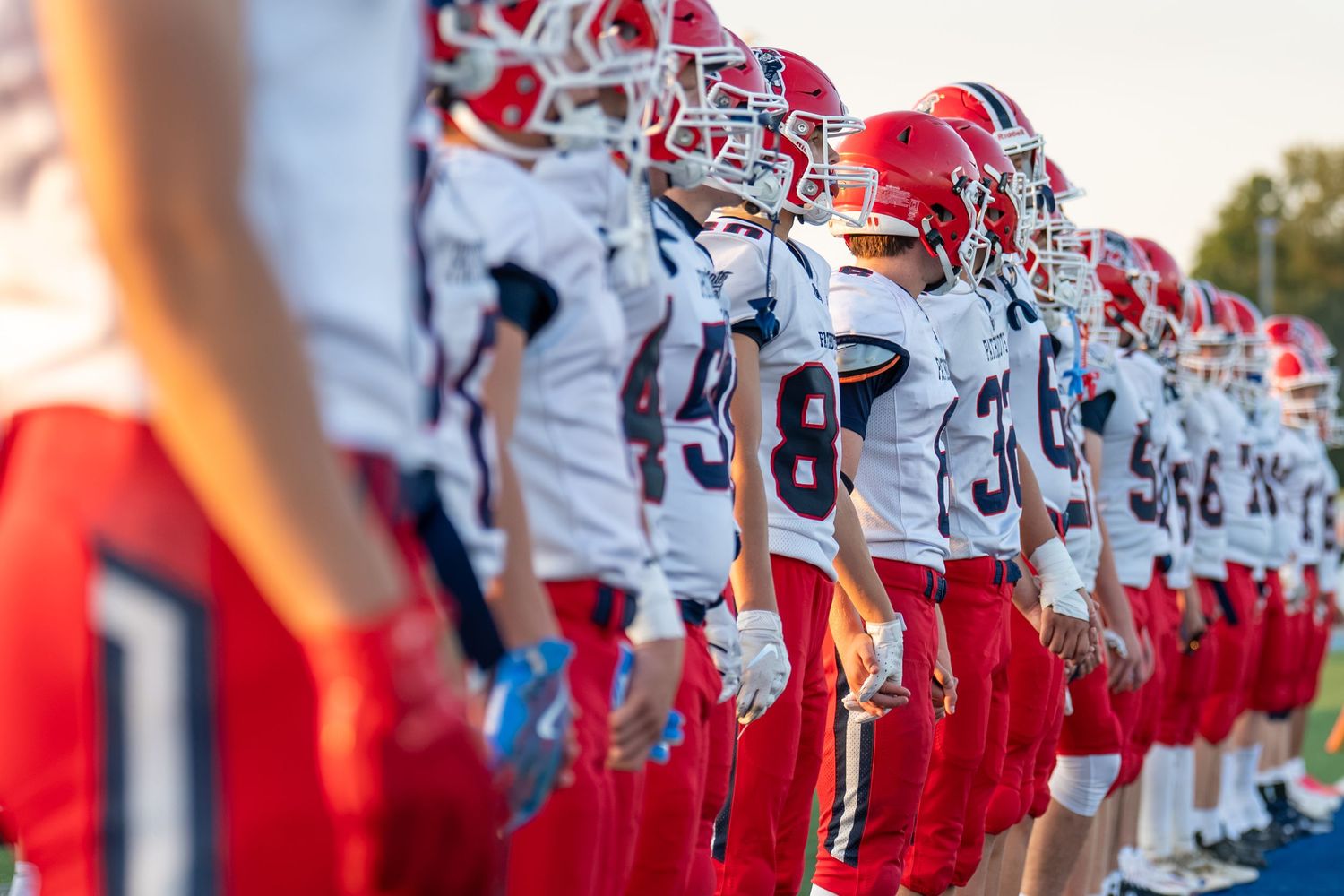 Youth football players in red and white uniforms line up on the sideline during a game.