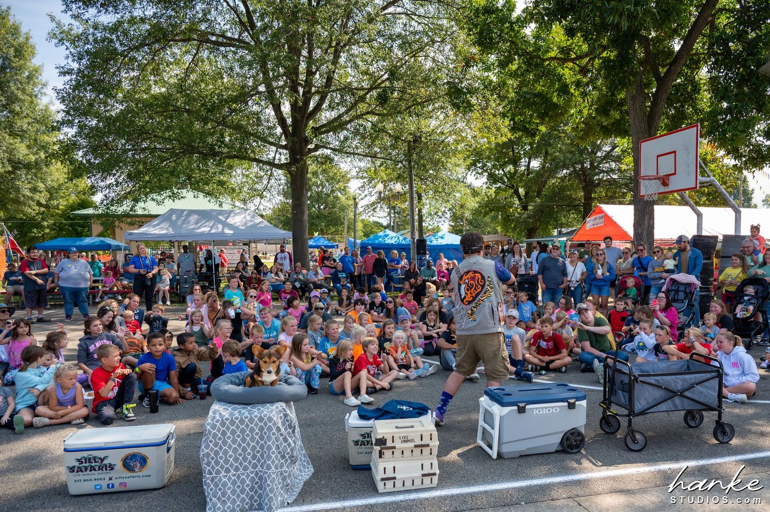 Large crowd of children and adults gather outdoors under trees for a community event presentation.