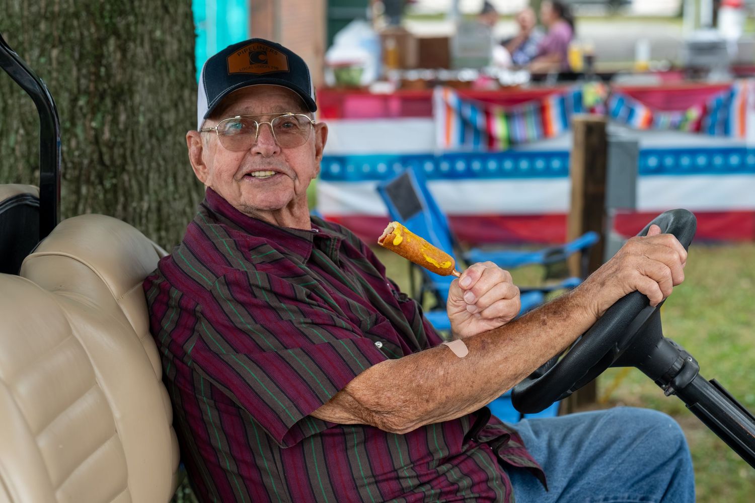 Elderly man in plaid shirt and cap smiles while driving a golf cart, holding a corn dog at an outdoor fair.