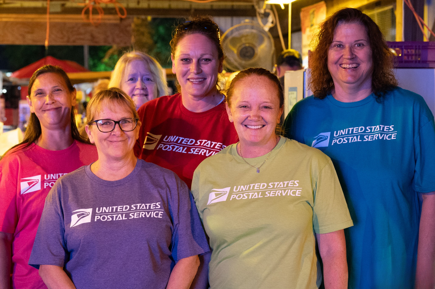 Six women in United States Postal Service t-shirts smile together for a group photo indoors.