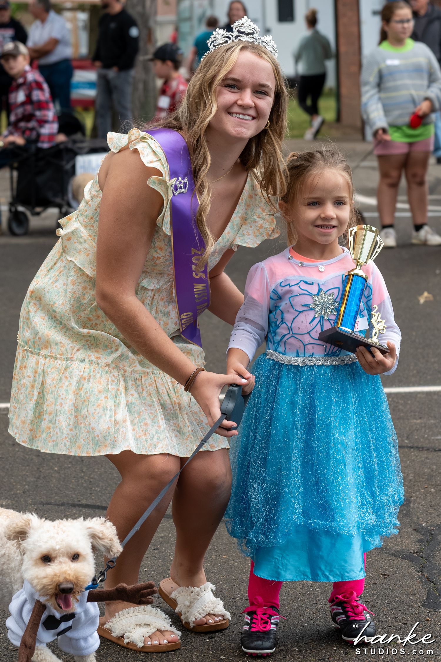 A teen beauty queen in a tiara and sash poses with a young girl holding a trophy and a small dressed-up dog at a parade.