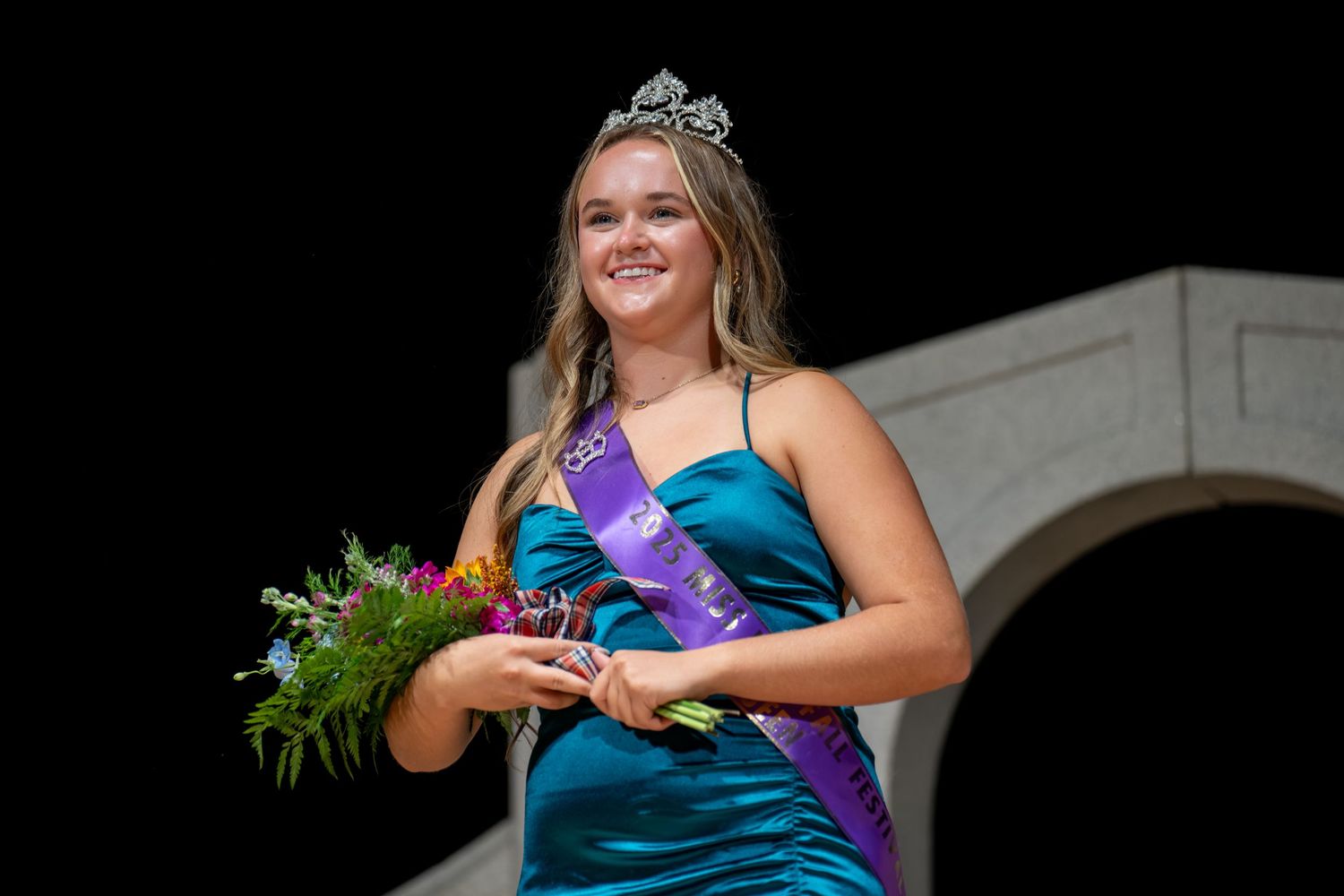 Young woman in teal gown wearing crown and purple sash holds bouquet at pageant competition.