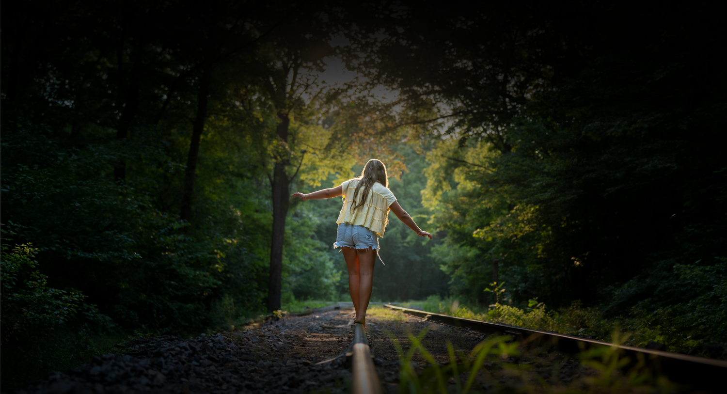 A young woman balances on railroad tracks in a lush green forest tunnel, arms outstretched, walking away from camera.
