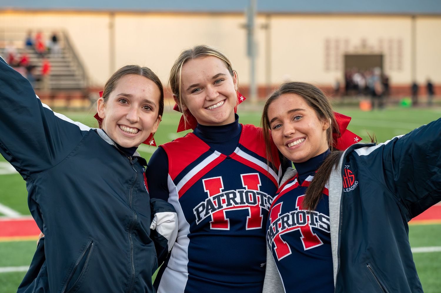 Three smiling teenage girls in Patriots cheerleading uniforms pose together on a football field.
