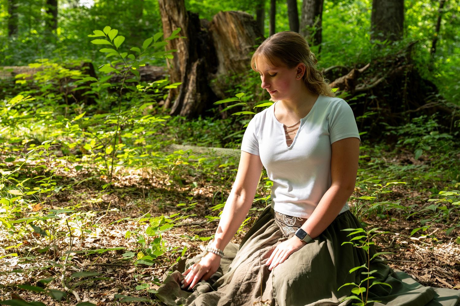 Young woman in white top and olive skirt kneels peacefully in sunlit woodland setting.