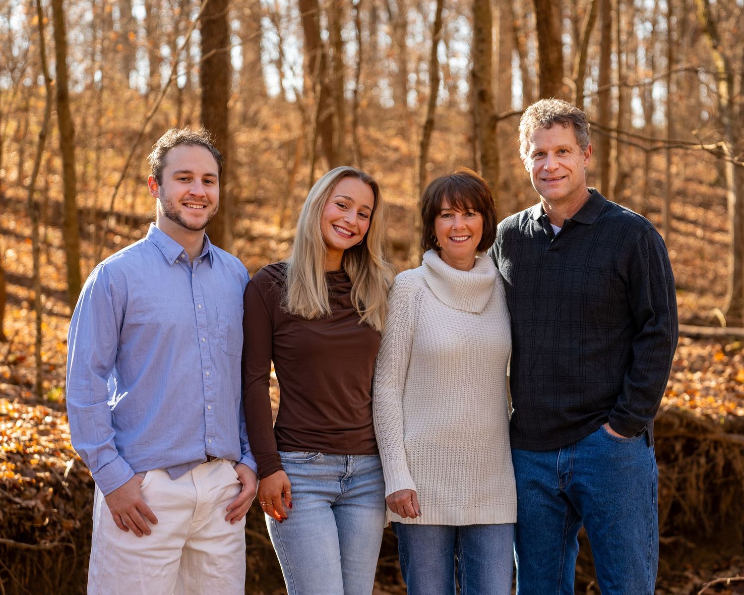 Family of four smiling outdoors in autumn woods, posing together for a fall portrait.