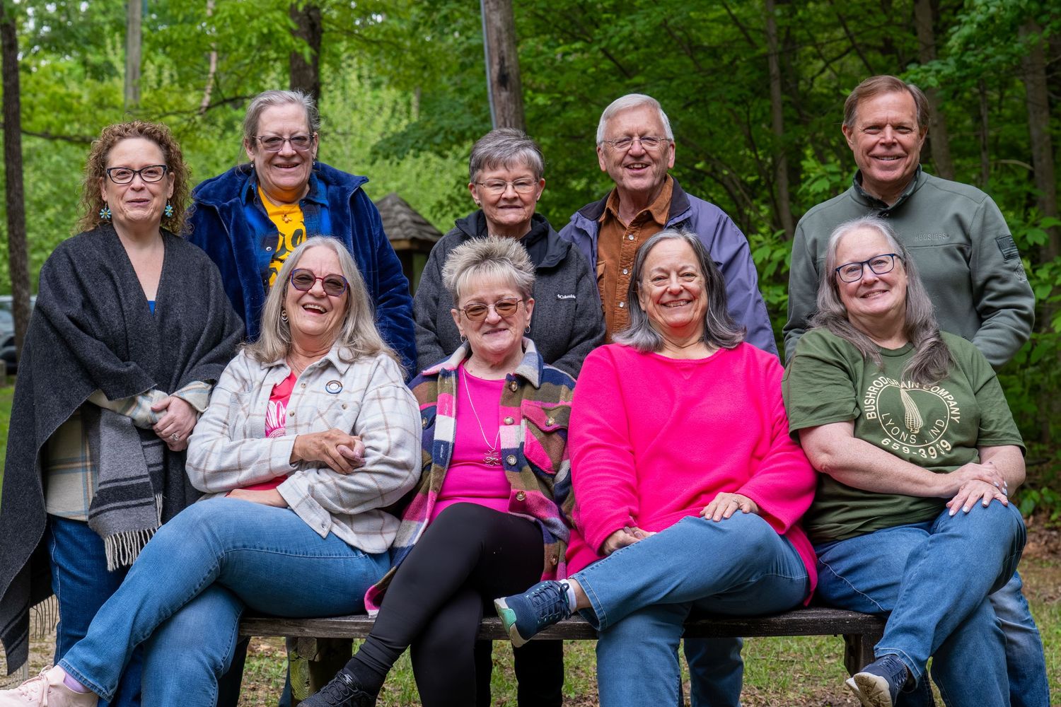 Group of nine older adults posing together outdoors on benches in a wooded park setting.